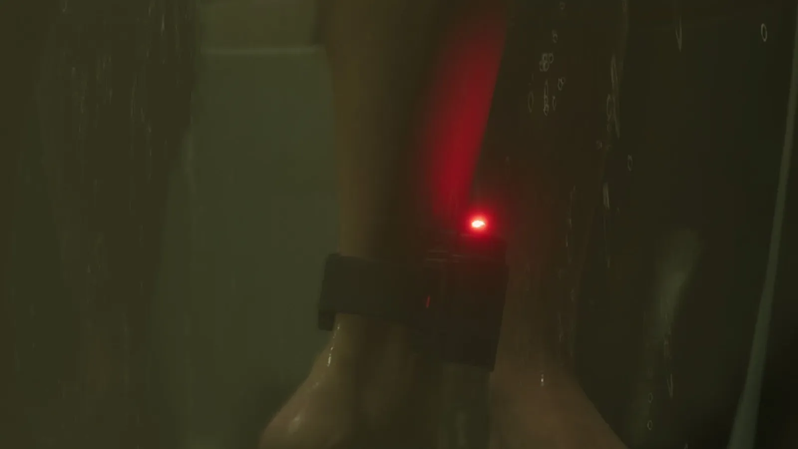 Close-up of a bare ankle in a dark shower with a black ankle monitor blinking a red light