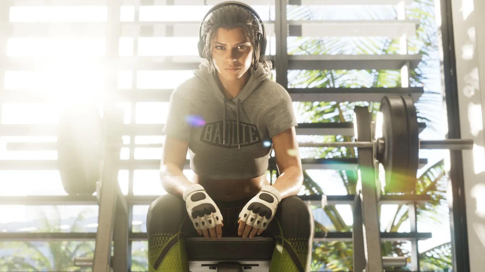Lucia, in headphones, a cropped hoodie and fingerless gloves, sits on a weight bench in a sunlit gym