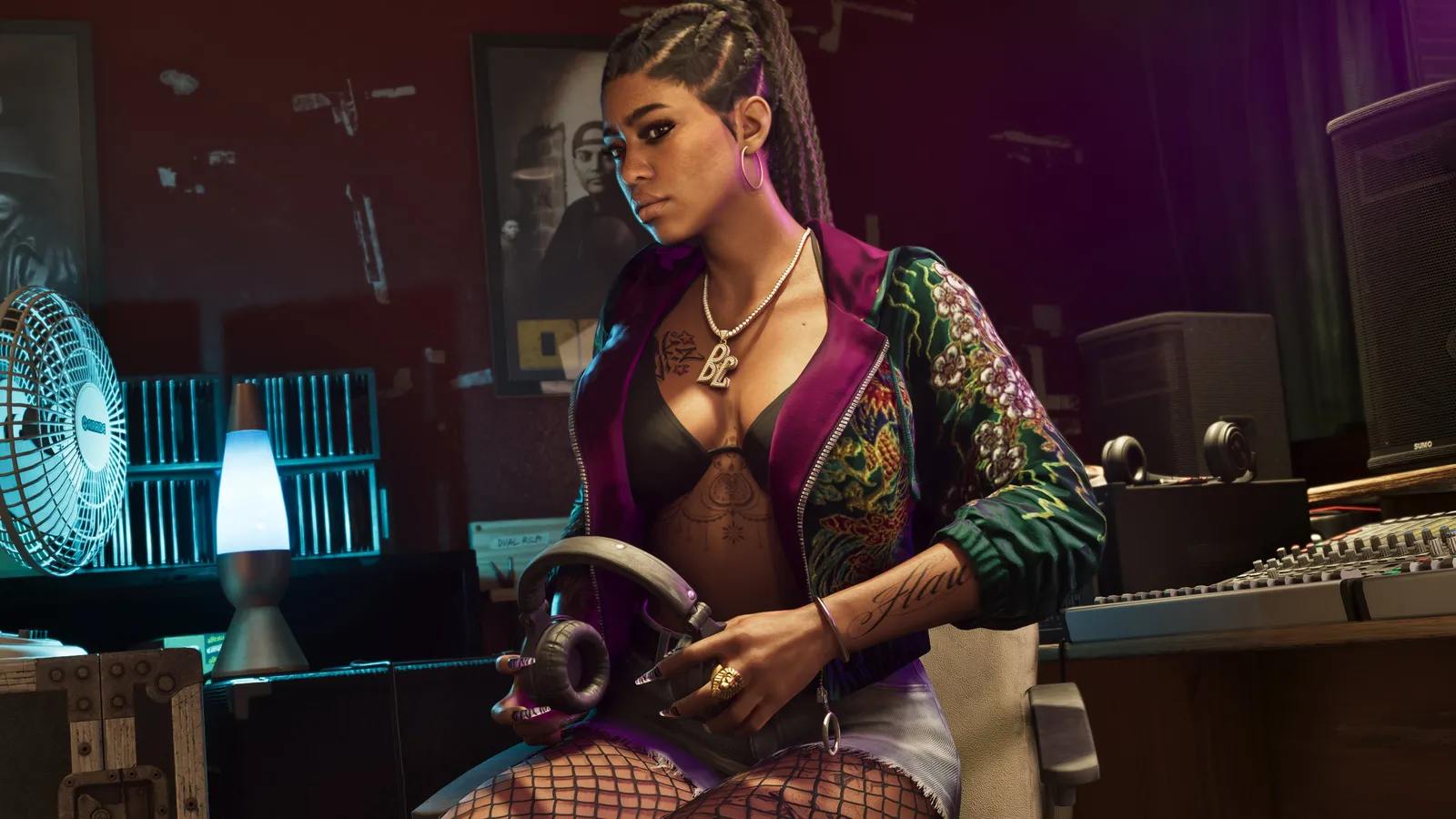 A member of Real Dimez in a recording studio, holding a pair of headphones beside a mixing desk, a desk fan and a lava lamp